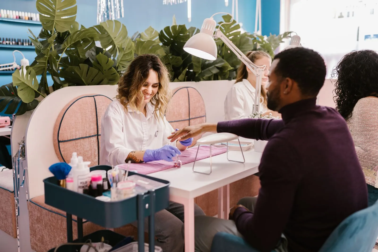 Manikür masasında çalışan nail artist ve müşterisi, arkada yeşil bitkiler